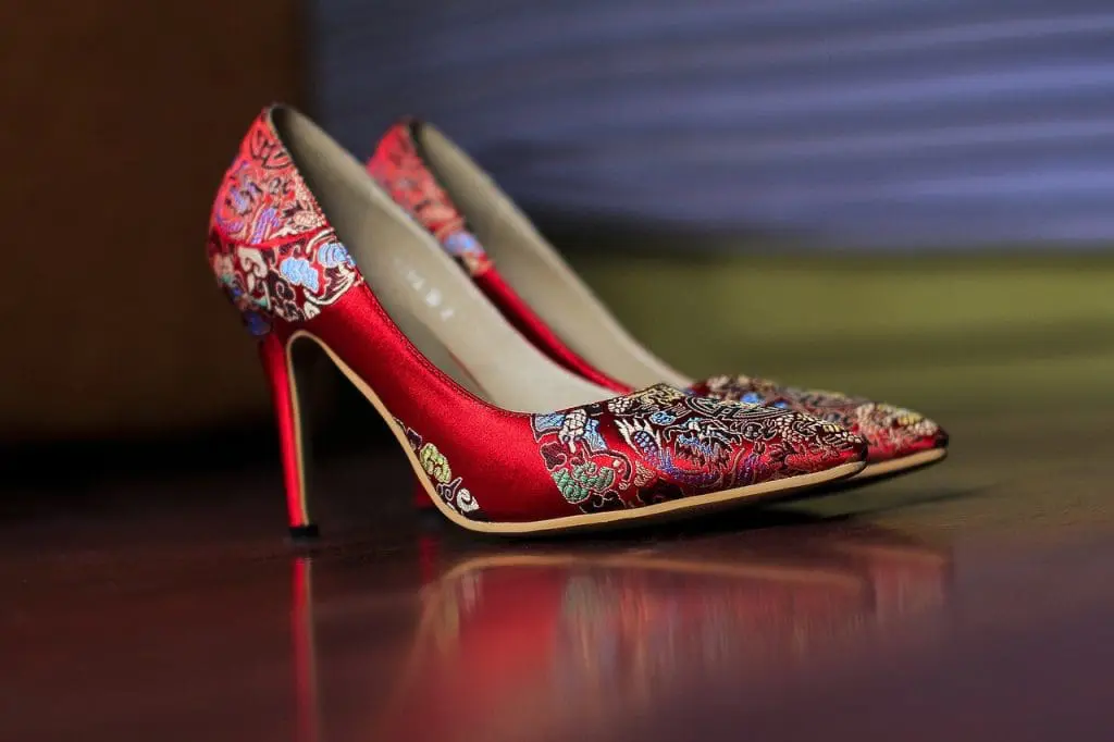 A pair of elegant high-heeled shoes with pointed toes, covered in shiny red fabric and intricate multicolored floral and paisley patterns, sits on a dark wooden floor—perfect for embracing your style through menopause with natural management.
