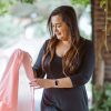 Mangla Sachdev, a woman with long brown hair, wearing a black dress and a smartwatch, holds a light pink sheer fabric. She is smiling and looking down at the fabric, with greenery and a brick structure softly blurred in the background.