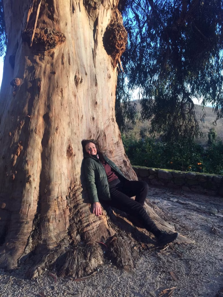 a person in a green jacket and black pants sits leaning back against the thick trunk of a large tree, smiling and relaxed. surrounded by greenery and sunlight, they radiate calm sensual energy as the tree’s textured bark glows in long shadows.
