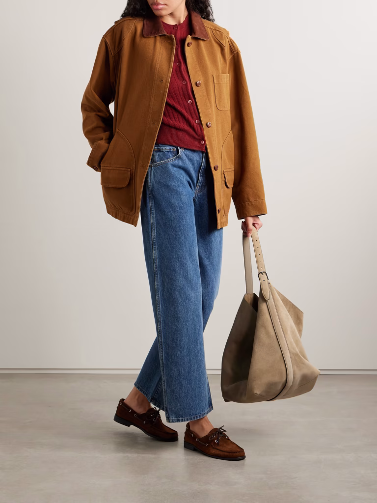a person stands indoors wearing a tan jacket over a red shirt, blue jeans, and brown loafers—an outfit that captures relaxed autumn winter 2025 style. they hold a large beige tote bag against a plain light wall and concrete floor.