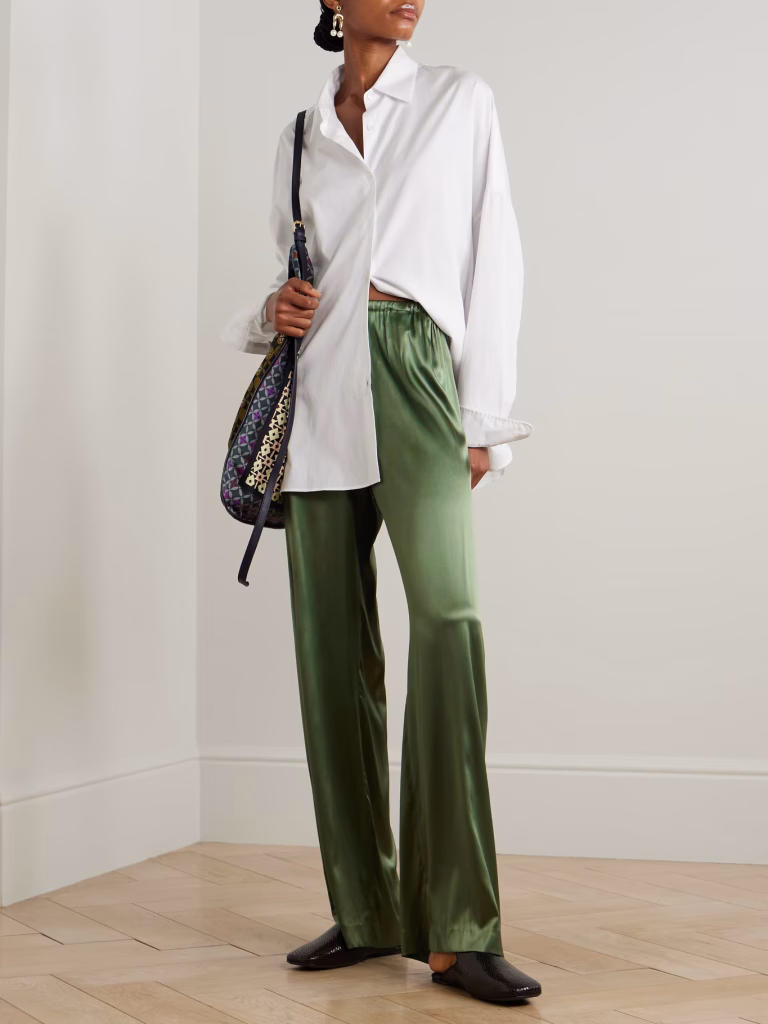 channeling summer trends, a person stands indoors in a loose white button down shirt, shiny green satin pants, and black shoes. they carry a patterned shoulder bag and wear earrings, set against light walls and wooden flooring with their head turned to the side.