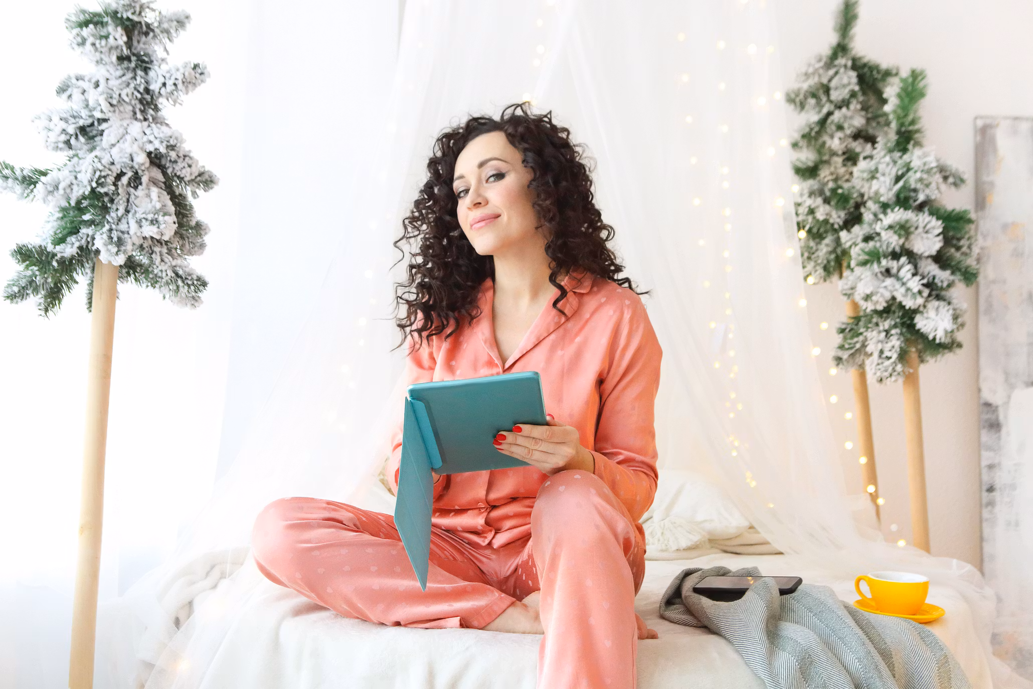 a woman with curly hair, in pink pajamas, sits cross legged on a bed holding a teal tablet, reading a romance novel during the Christmas holidays. the room, glowing with string lights and snowy trees, is perfect for diving into must read christmas books. a yellow cup and gray scarf rest beside her.