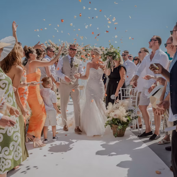 a bride and groom walk arm in arm down an outdoor aisle at oceana weddings spain, smiling joyfully as guests in summer attire toss flower petals. the mediterranean sun shines as everyone celebrates this beautiful destination wedding venue.