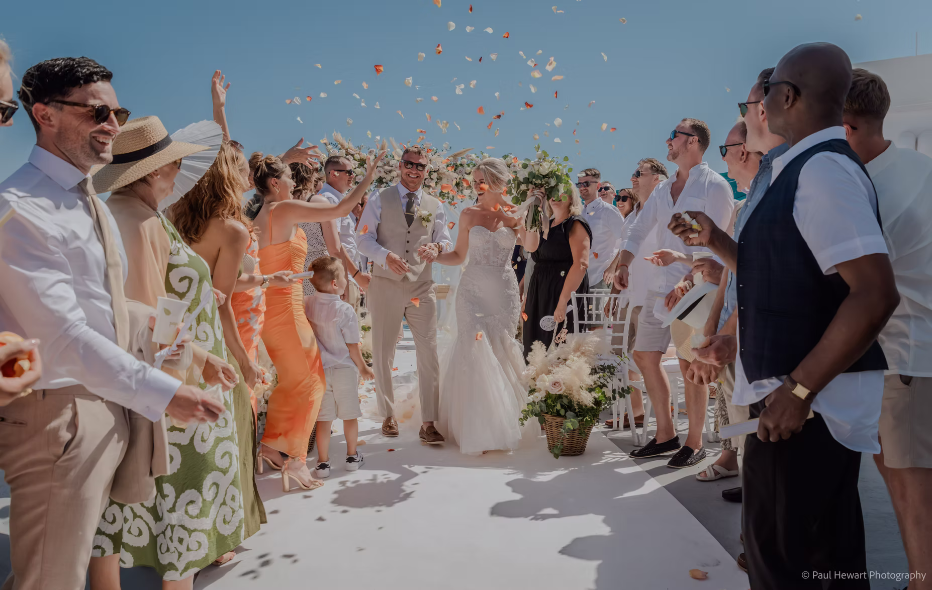 a bride and groom walk arm in arm down an outdoor aisle at oceana weddings spain, smiling joyfully as guests in summer attire toss flower petals. the mediterranean sun shines as everyone celebrates this beautiful destination wedding venue.