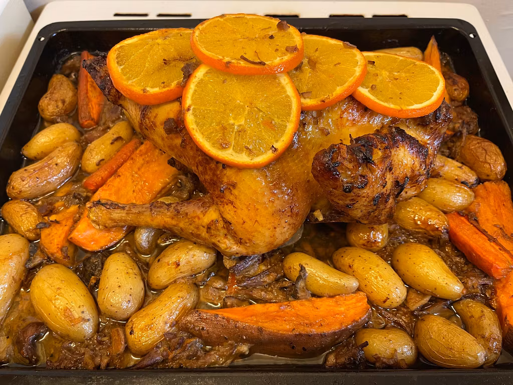a whole roasted chicken topped with orange slices sits in a baking tray surrounded by fingerling potatoes and carrots, all coated in a golden brown glaze—perfect for bringing tradition to your festive table with this rustic homemade meal. A whole roasted chicken topped with orange slices sits in a baking tray surrounded by fingerling potatoes and carrots, all coated in a golden-brown glaze—perfect for bringing tradition to your festive table with this rustic homemade meal.