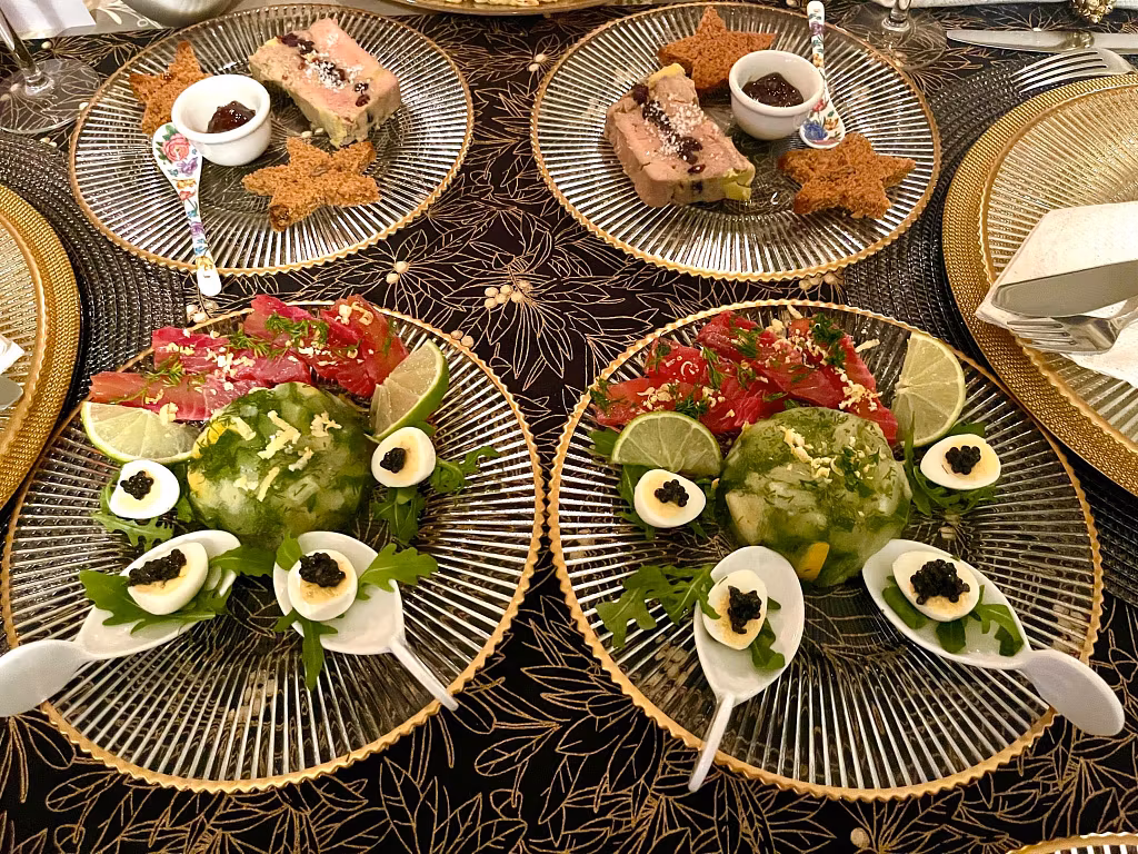 Two elegant plates with green aspic salads, festive starters garnished with lime slices, parsley, and deviled eggs topped with caviar. Nearby plates hold pâté slices with dark fruit jelly and star-shaped crackers on gold-rimmed glass plates.