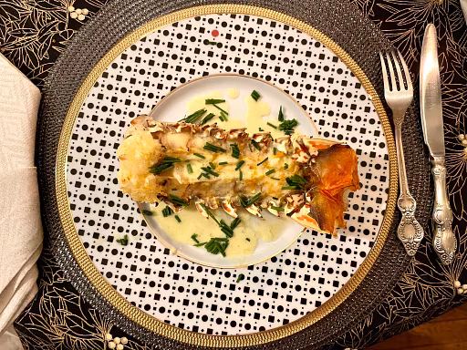 A plated lobster tail topped with chopped herbs and creamy sauce sits on a white plate with a black dotted pattern, evoking the sea, while ornate silverware and a white napkin complete this festive table set on a black floral tablecloth.