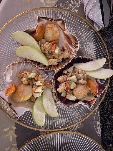 Three scallop shells on a glass plate evoke the sea, each filled with scallops, diced vegetables, and citrusy green apple slices. The gold-rimmed plate rests on a festive tablecloth with a folded napkin in the upper right corner.
