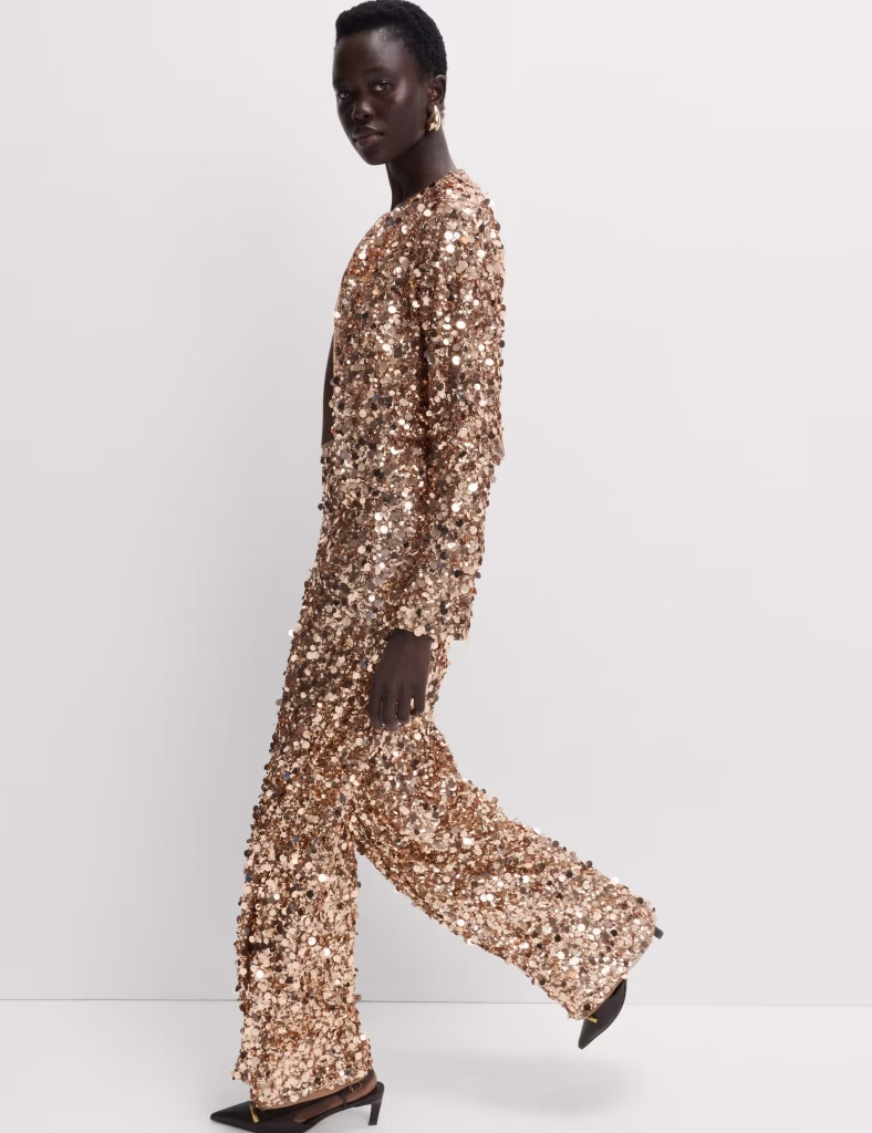 A person with short hair walks in profile, wearing a shimmering gold sequin-covered long-sleeve top and matching wide-leg pants—perfect for Christmas styling. They complete the look with pointed black heels and gold earrings against a plain off-white background.