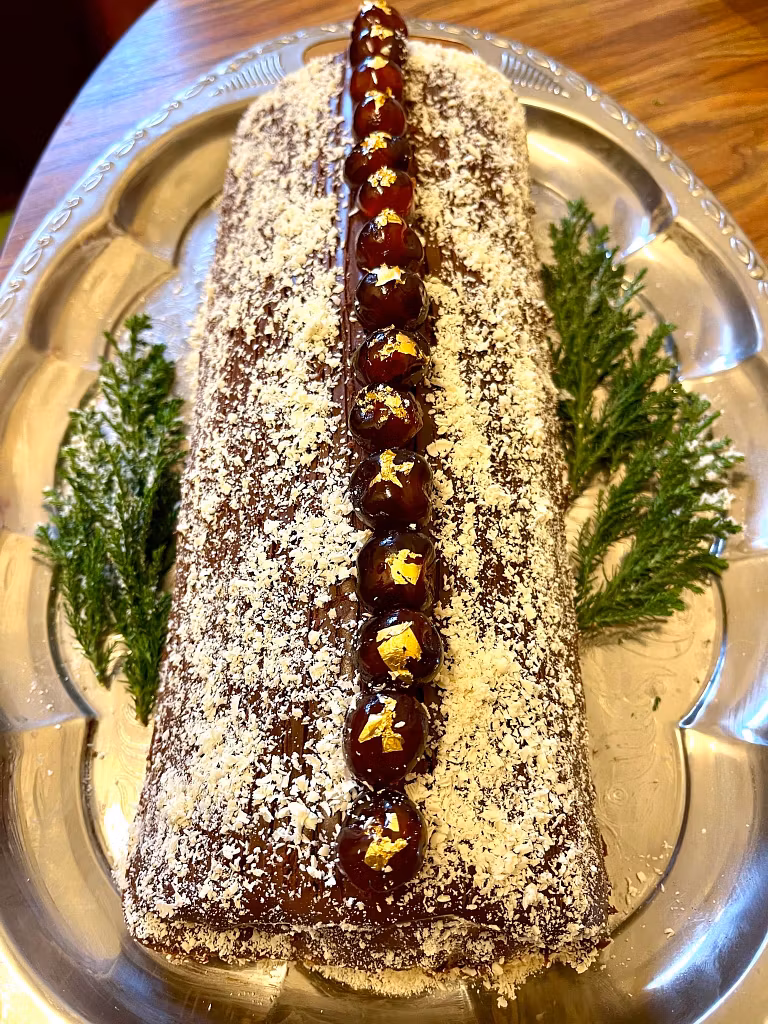 Tradition Reimagined for the Festive Table A reimagined tradition, this rectangular chocolate Yule log cake sits on a silver tray, dusted with coconut flakes. Glossy cherries topped with gold leaf line the center, greenery decorates each side, and it graces a festive table.