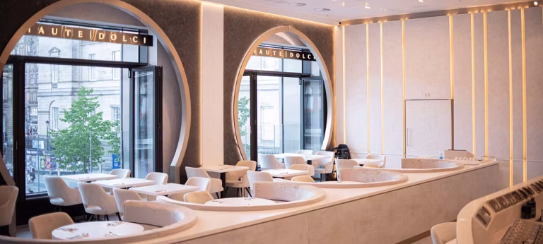 A modern, elegant restaurant interior with large arched windows, round white tables, and cushioned white chairs. Soft yellow lighting and curved features create a sophisticated atmosphere—perfect for enjoying HAUTE DOLCI’s divine desserts.