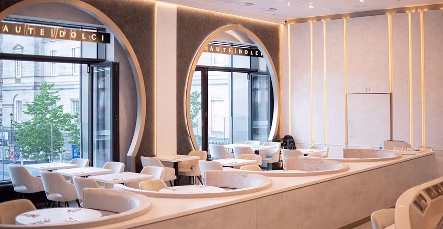 A modern, elegant restaurant interior with large arched windows, round white tables, and cushioned white chairs. Soft yellow lighting and curved features create a sophisticated atmosphere—perfect for enjoying HAUTE DOLCI’s divine desserts.