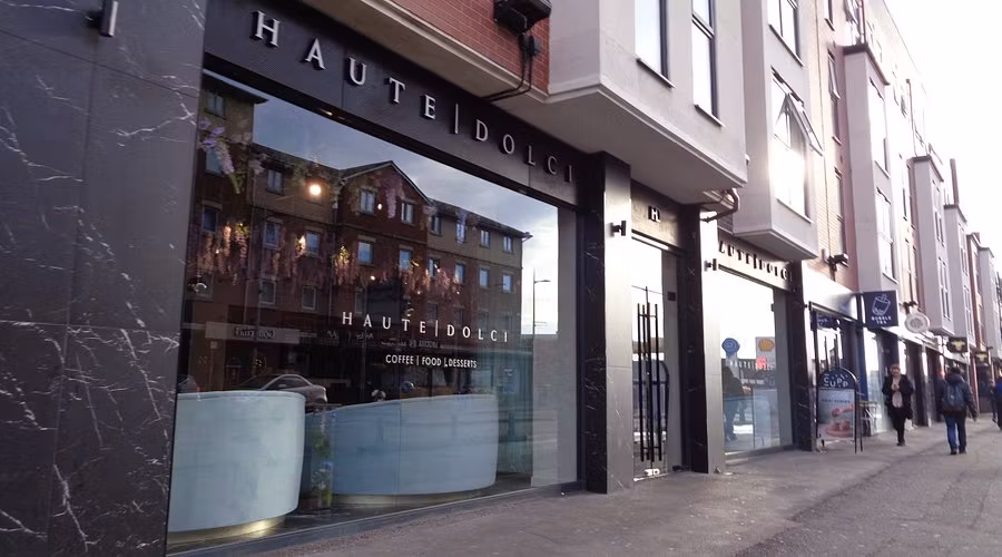 street view of the modern café haute dolci, with large glass windows reflecting nearby buildings. the stylish black exterior and white signage hint at the divine desserts inside, as pedestrians stroll past neighboring shops on a cloudy day. Street view of the modern café HAUTE DOLCI, with large glass windows reflecting nearby buildings. The stylish black exterior and white signage hint at the divine desserts inside, as pedestrians stroll past neighboring shops on a cloudy day.