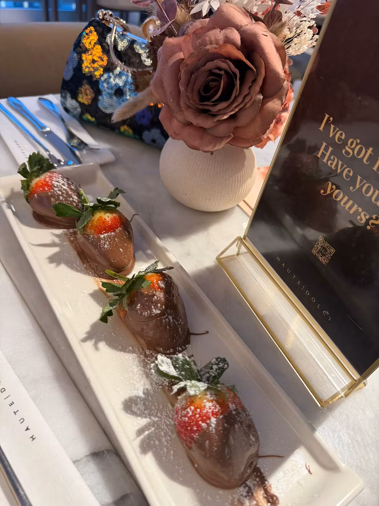 a rectangular white plate holds four strawberries dipped in chocolate—one of haute dolci’s divine desserts—garnished with powdered sugar and mint leaves. the plate sits on a marble table beside a mauve artificial rose and a chic floral handbag. A rectangular white plate holds four strawberries dipped in chocolate—one of HAUTE DOLCI’s divine desserts—garnished with powdered sugar and mint leaves. The plate sits on a marble table beside a mauve artificial rose and a chic floral handbag.