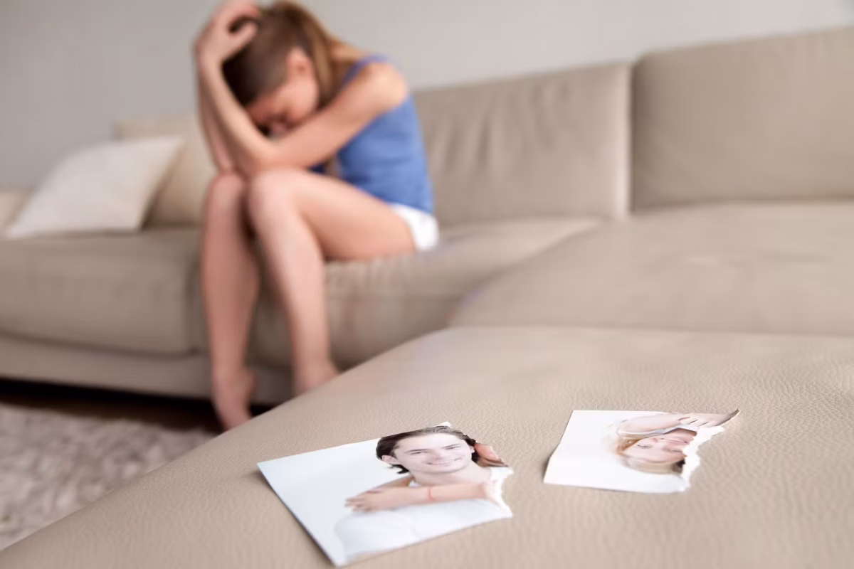A woman sits on a beige couch with her head in her hands, appearing upset. In the foreground, a torn photograph of a smiling couple lies on the couch, symbolizing heartbreak and a relationship dilemma. The background is blurred, focusing on the photo.