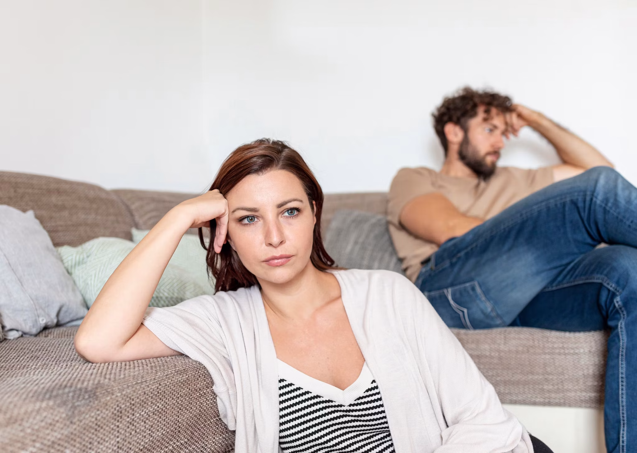 A woman sits on a couch, looking serious and lost in thought. In the background, a man gazes away, both caught in a relationship dilemma. Their distance suggests they’re quietly wondering, “Should I Stay or Should I Go?”.