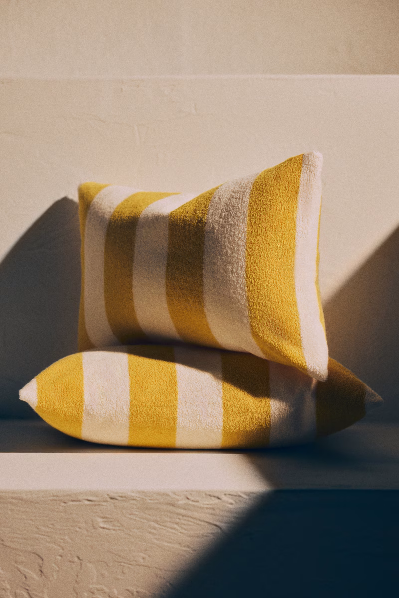 Two rectangular pillows with thick, horizontal yellow and white stripes—perfect for spring trends—are stacked on a textured, light-colored surface. Soft lighting highlights their plush texture, creating a warm and inviting high street-inspired scene.
