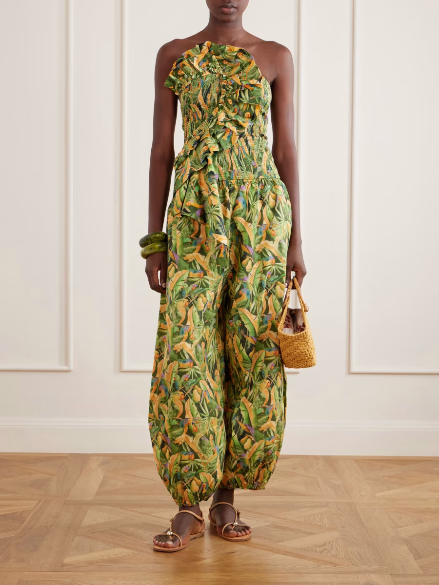 A person stands indoors wearing a sleeveless, ruffled green and yellow botanical-print jumpsuit with balloon trousers for a voluminous silhouette. They hold a woven straw handbag, and wear gold sandals and a chunky green bracelet against a paneled wall.