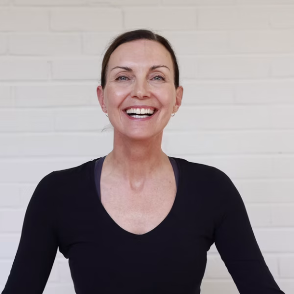Home A woman with dark hair pulled back smiles at the camera. She wears a fitted black top and stands against a plain white brick wall backdrop. The lighting is bright and even, highlighting her cheerful expression—perfectly capturing the positive energy she brings to every yoga routine.