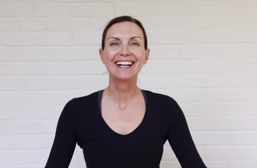 A woman with dark hair pulled back smiles at the camera. She wears a fitted black top and stands against a plain white brick wall backdrop. The lighting is bright and even, highlighting her cheerful expression—perfectly capturing the positive energy she brings to every yoga routine.